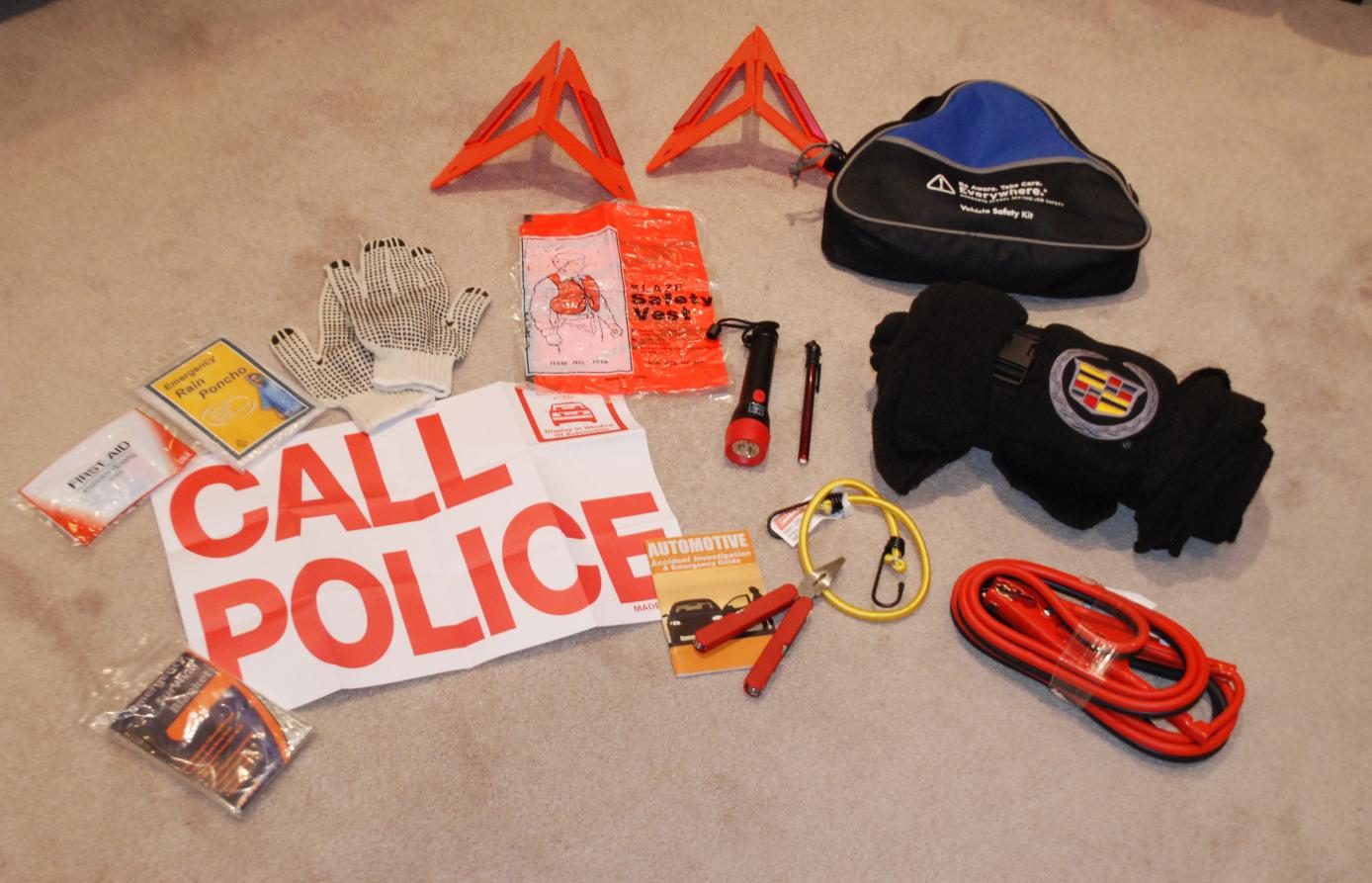 An example of a pre-made emergency kit. Items included are jumper cables, a bungee cord, a multipurpose tool, a blanket, a safety vest, a pair of gloves, reflective cones, a “Call Police” sign, a small booklet for car care, a poncho, a first aid kit, and another emergency blanket.