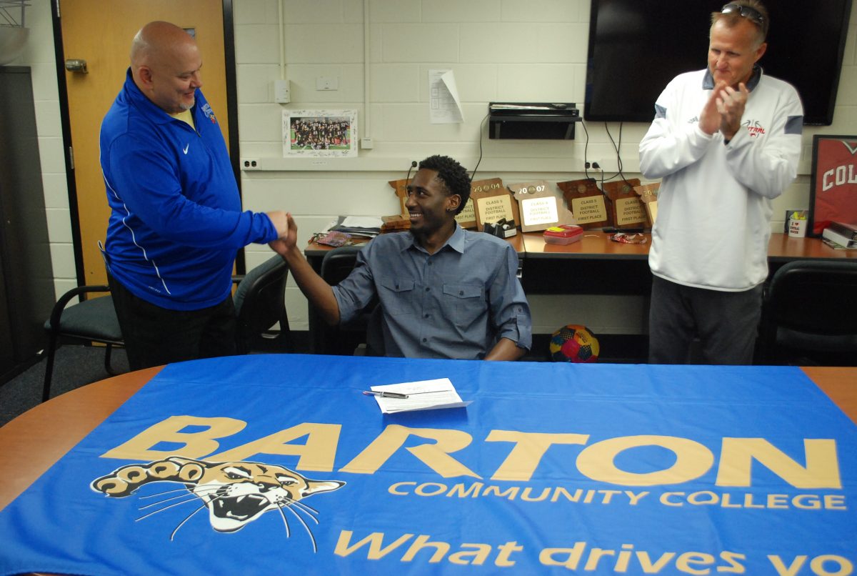 Senior Jarrett Cox-Bradley shakes hands with his new coach Craig Fletchall as his high school coach Rick Kirby applauds in the back. Cox-Bradley signed to Barton on Friday, April 10. Photo by Matthew Gibbs. 